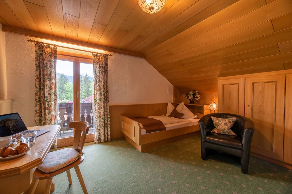 A cozy room with a wooden ceiling, a bed with a brown blanket, a chair, a table with croissants, and a laptop.