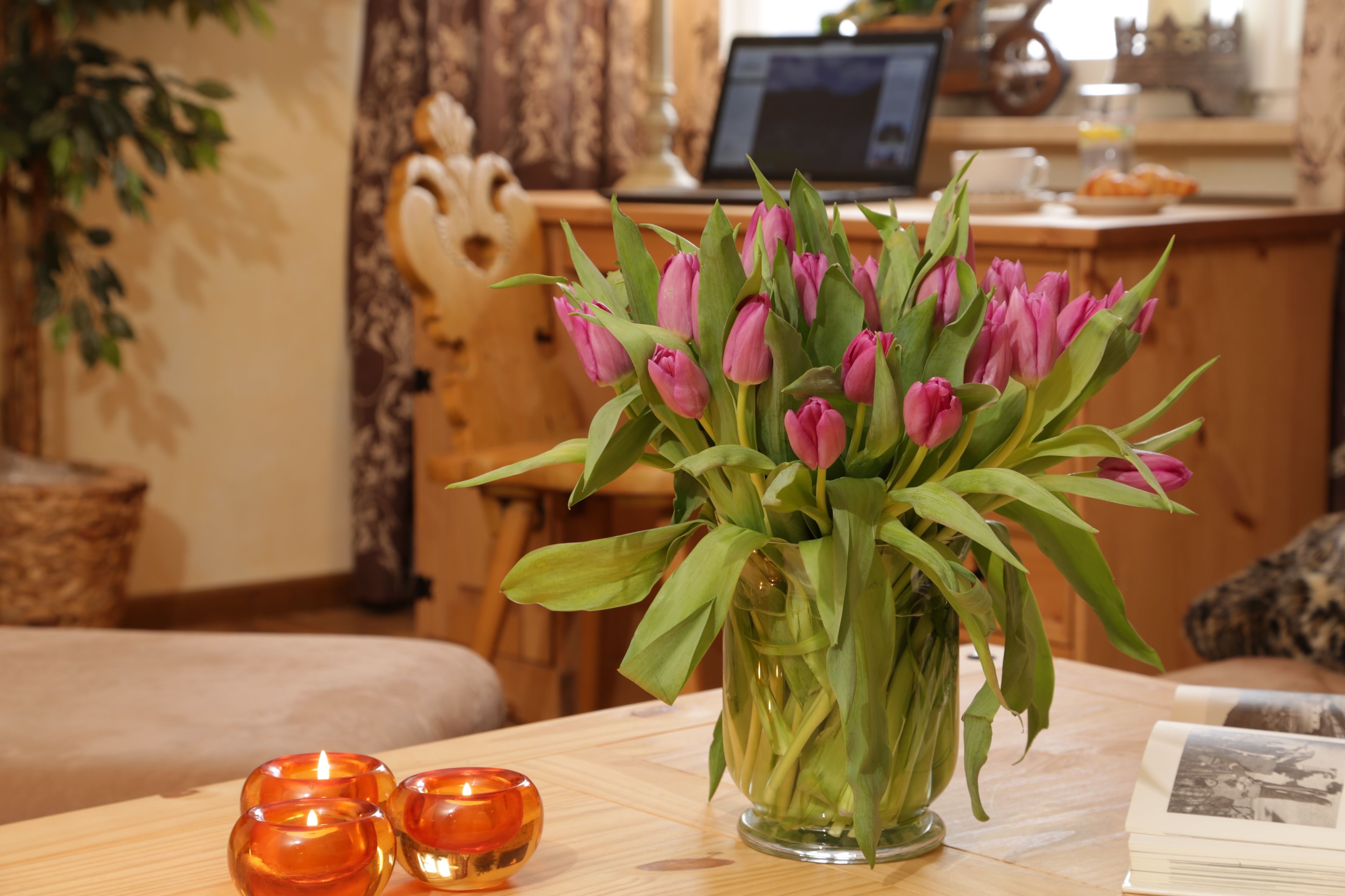 A wooden table with a vase of pink tulips, two orange candle holders, and a book.