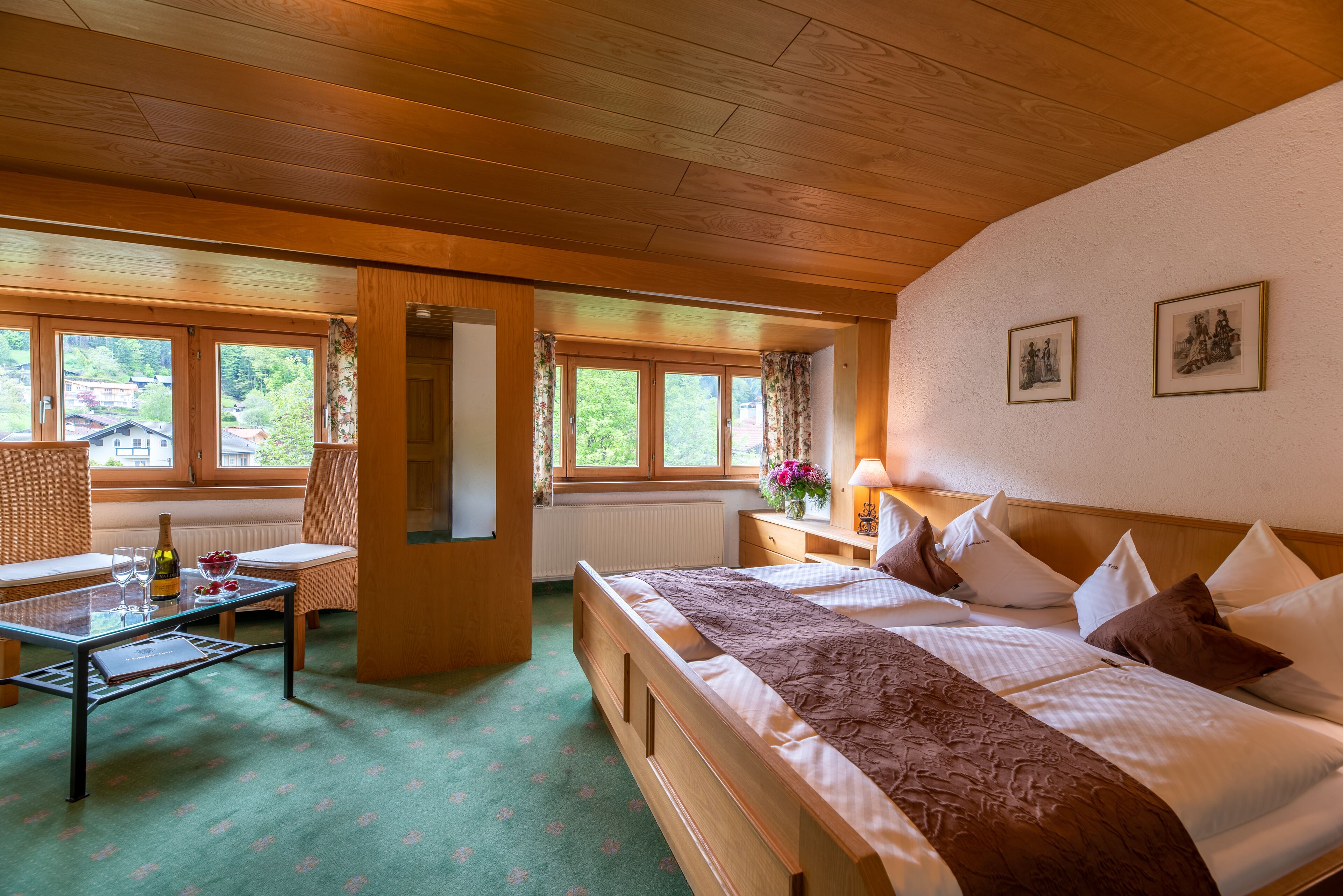 A hotel room with a wooden ceiling, a large bed with a brown bedspread, a glass table with a bottle and glasses, a chair, and a window with curtains.