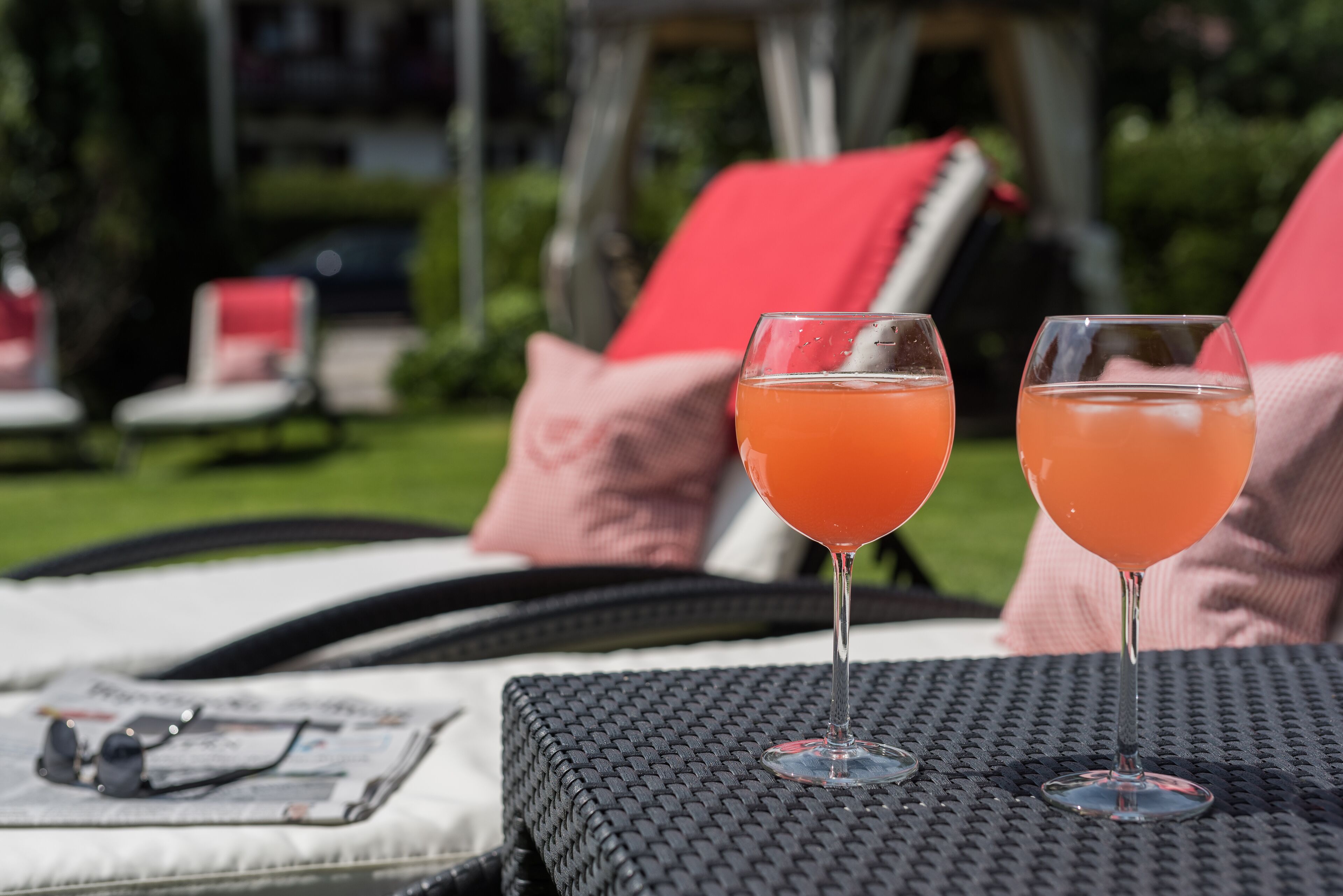 Dos vasos de bebida de naranja sobre una mesa, junto a un periódico, en un área de estar al aire libre con cojines rojos y una silla de mimbre.