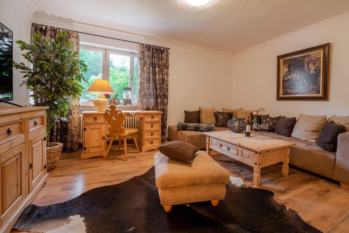 A cozy living room with a wooden coffee table, a sofa, a small armchair, and a dining table with chairs. There is a large potted plant, a lamp, and a painting on the wall.