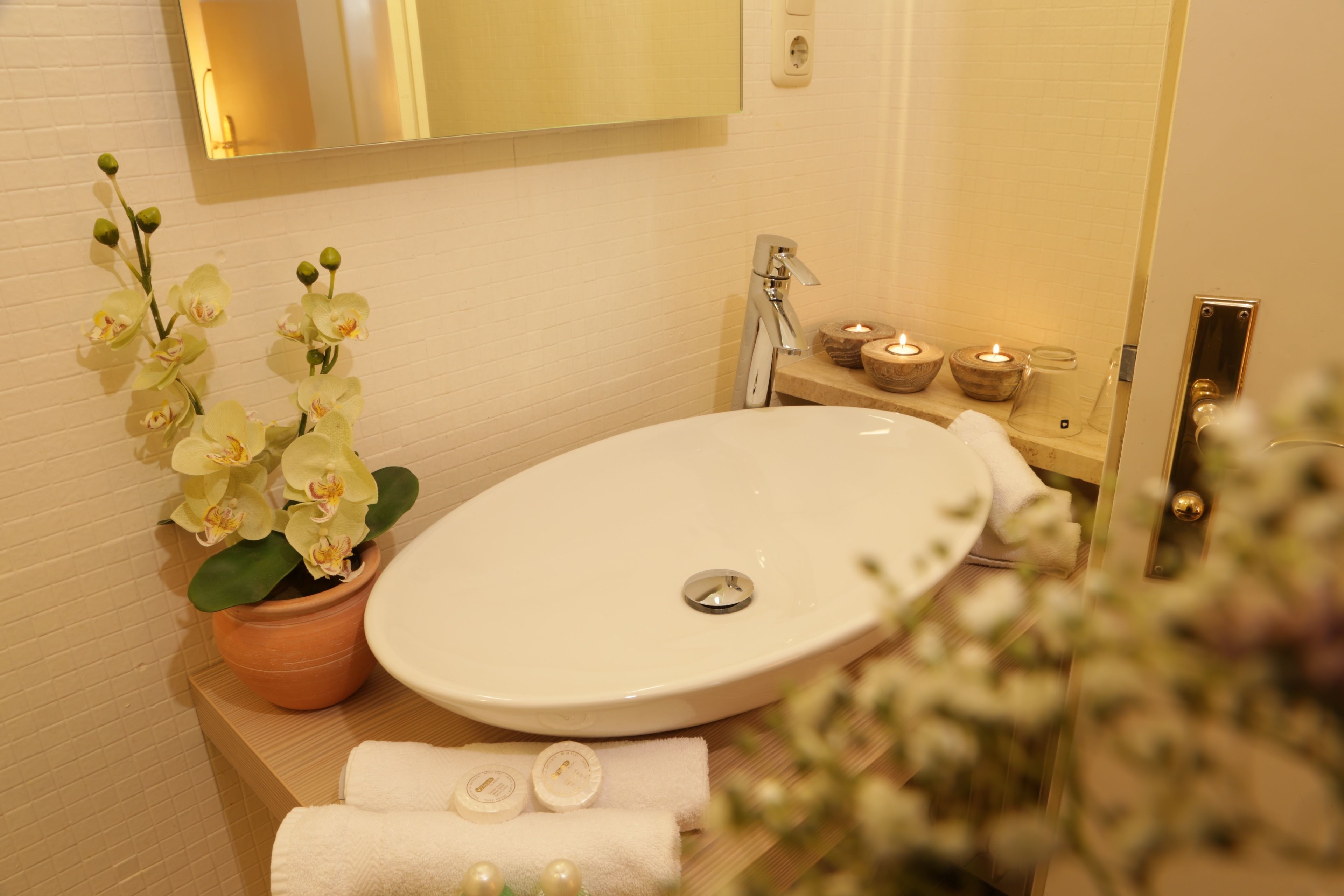 Un baño con lavamanos blanco, espejo, orquídea en maceta y un estante pequeño con velas.