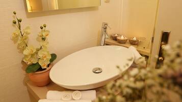 Un baño con lavamanos blanco, espejo, orquídea en maceta y un estante pequeño con velas.