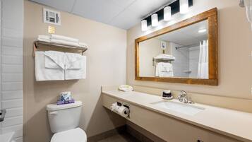 Une salle de bain équipée de toilettes, d’un lavabo et d’un grand miroir. Des serviettes sont pliées et empilées sur une étagère au-dessus des toilettes, et on aperçoit un rideau de douche dans le reflet du miroir.