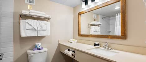 A bathroom with a toilet, sink, and a large mirror. There are towels stacked on a shelf above the toilet and a shower curtain visible in the mirror reflection.