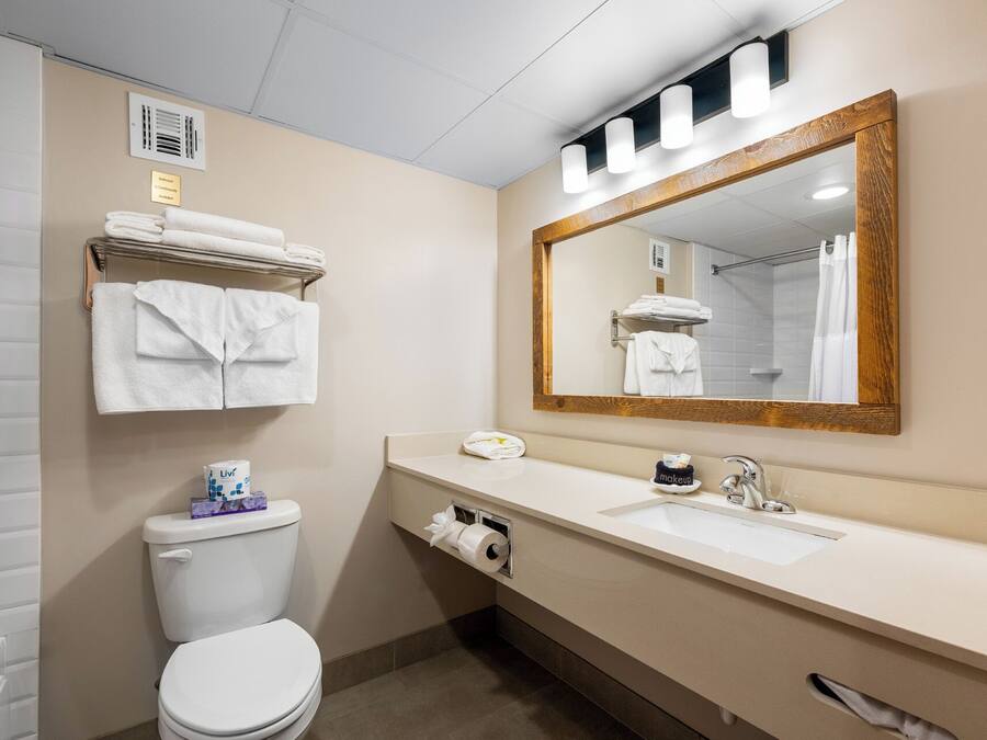 Un baño con inodoro, lavabo y un espejo grande. Hay toallas apiladas en un estante sobre el inodoro y se ve una cortina de ducha en el reflejo del espejo.