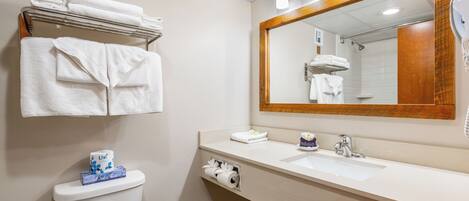 A bathroom with a toilet, sink, and mirror. There are towels on a rack and a box of toilet paper on the toilet.