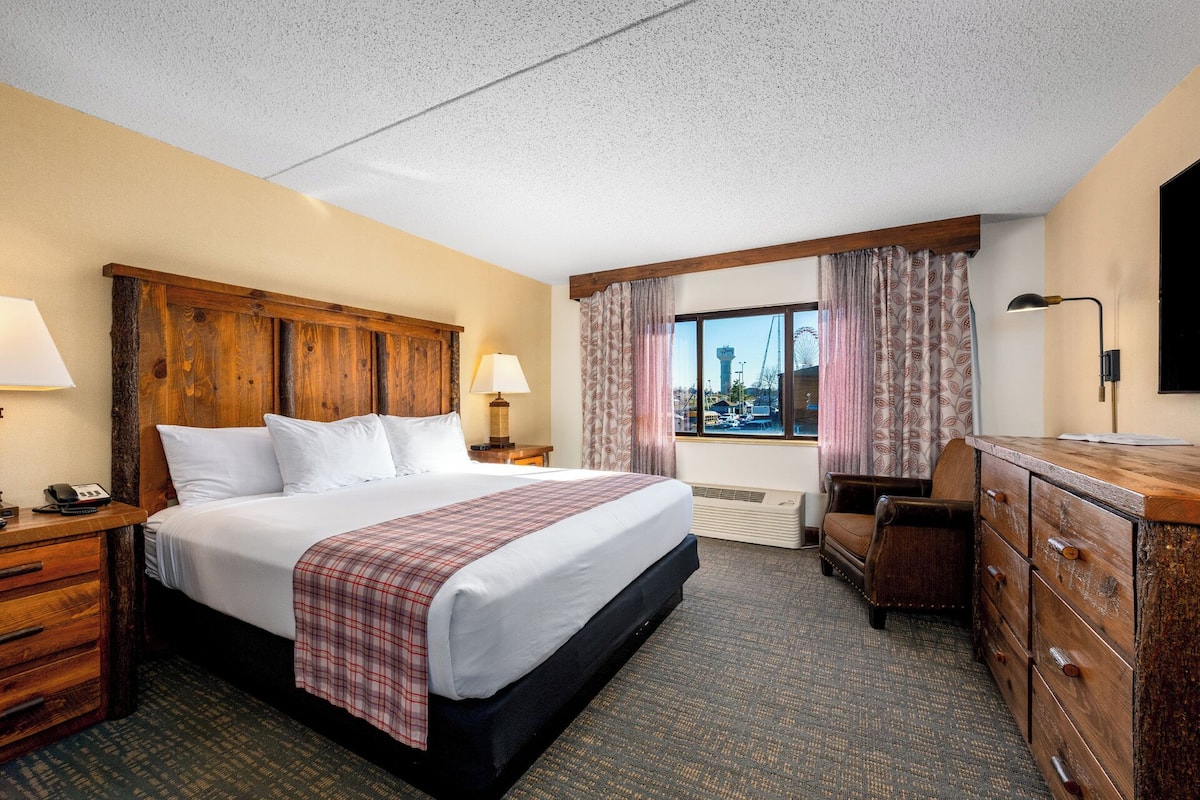 A hotel room with a large bed, a desk, a dresser, and a window with a view of a cityscape.