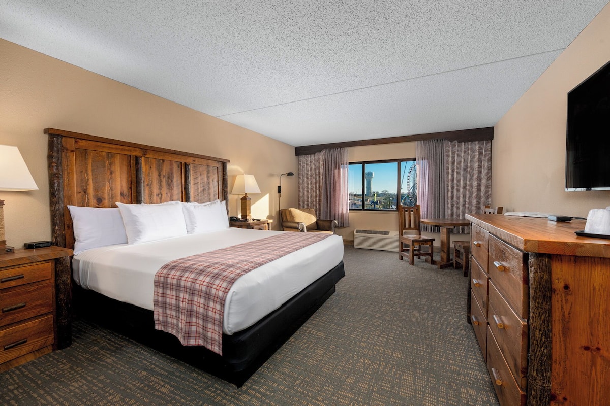 A hotel room with a large bed, a wooden headboard, a desk with a chair, a television, and a window with curtains.