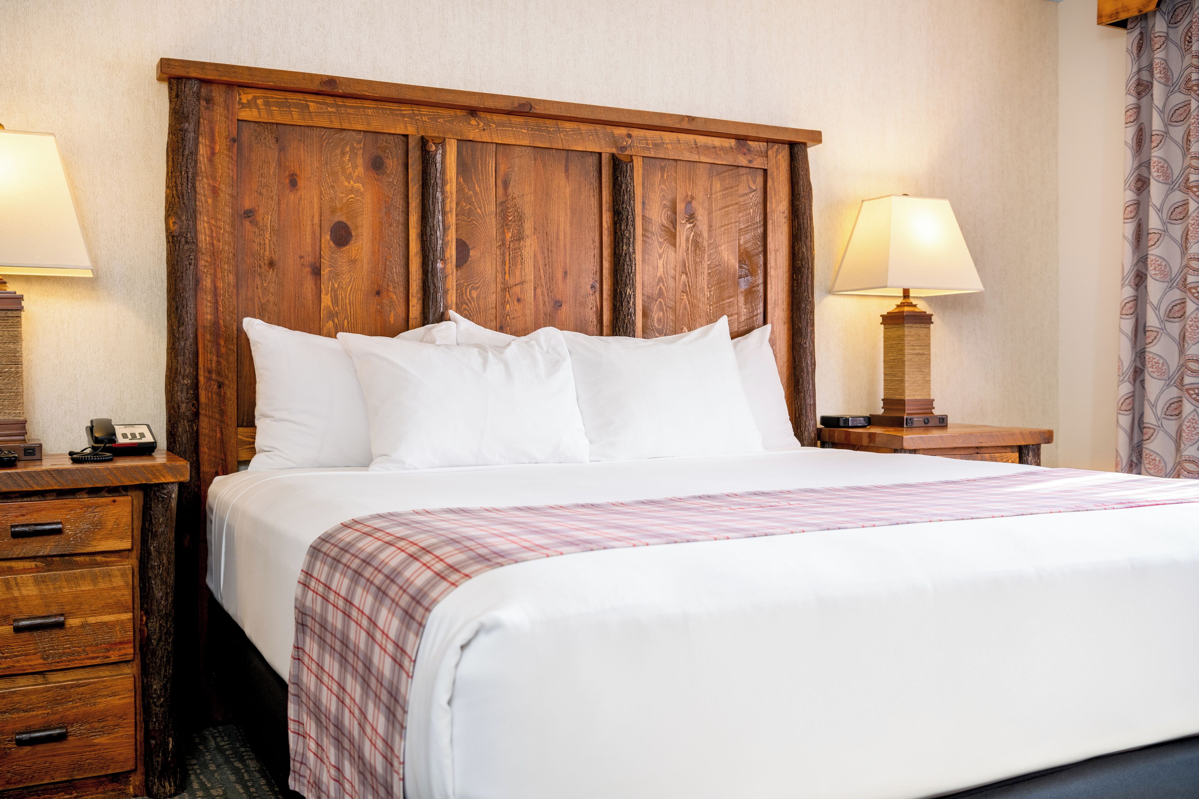 Una cama bien tendida con sábanas blancas y una manta escocesa, flanqueada por mesitas de noche de madera con lámparas, en una habitación con cabecera de madera.