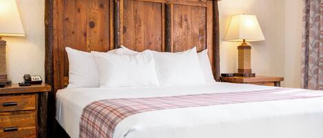 A neatly made bed with white linens and a plaid blanket, flanked by wooden nightstands with lamps, in a room with a wooden headboard.
