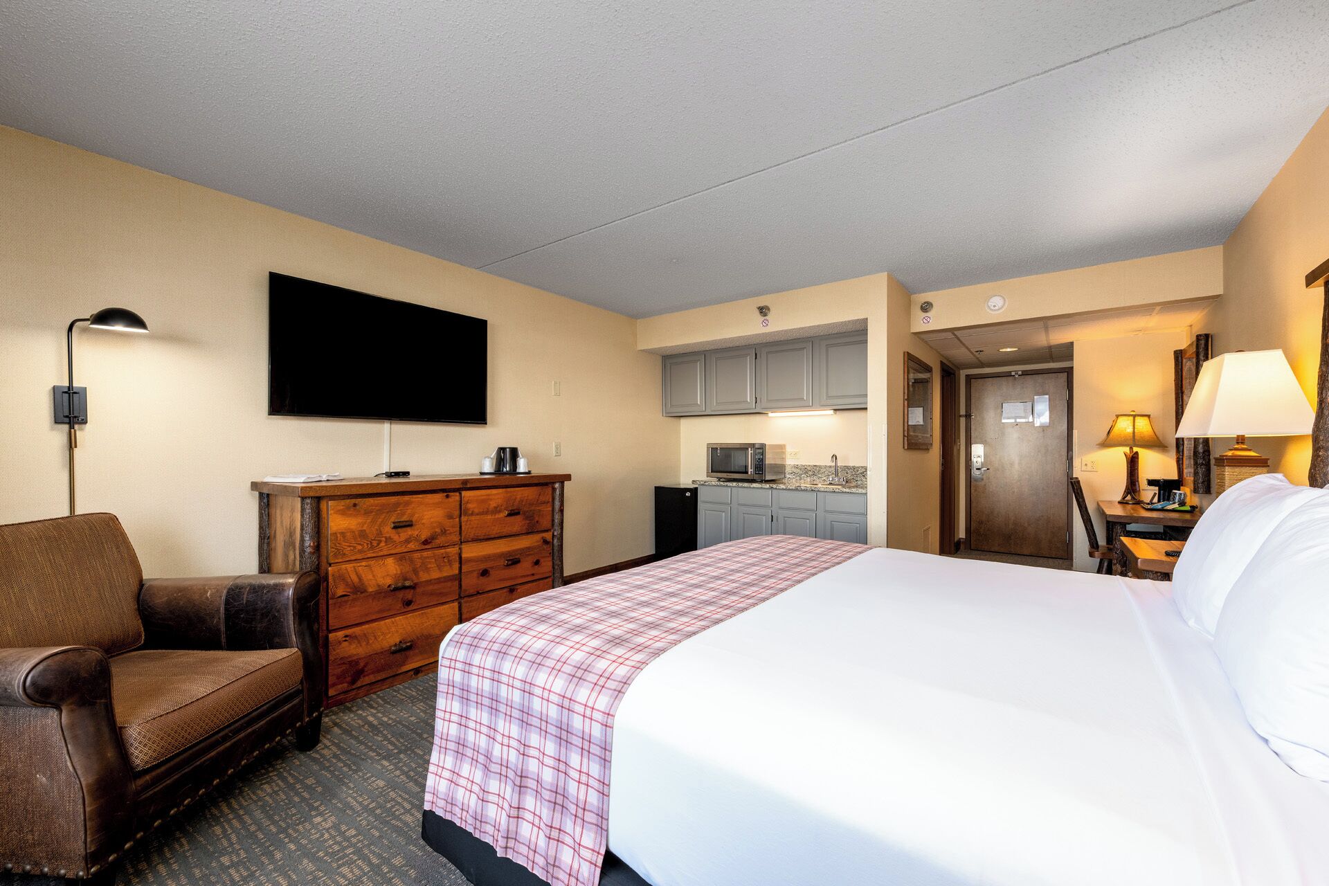 A hotel room with a bed, a dresser, a TV, a desk with a lamp, and a kitchenette.