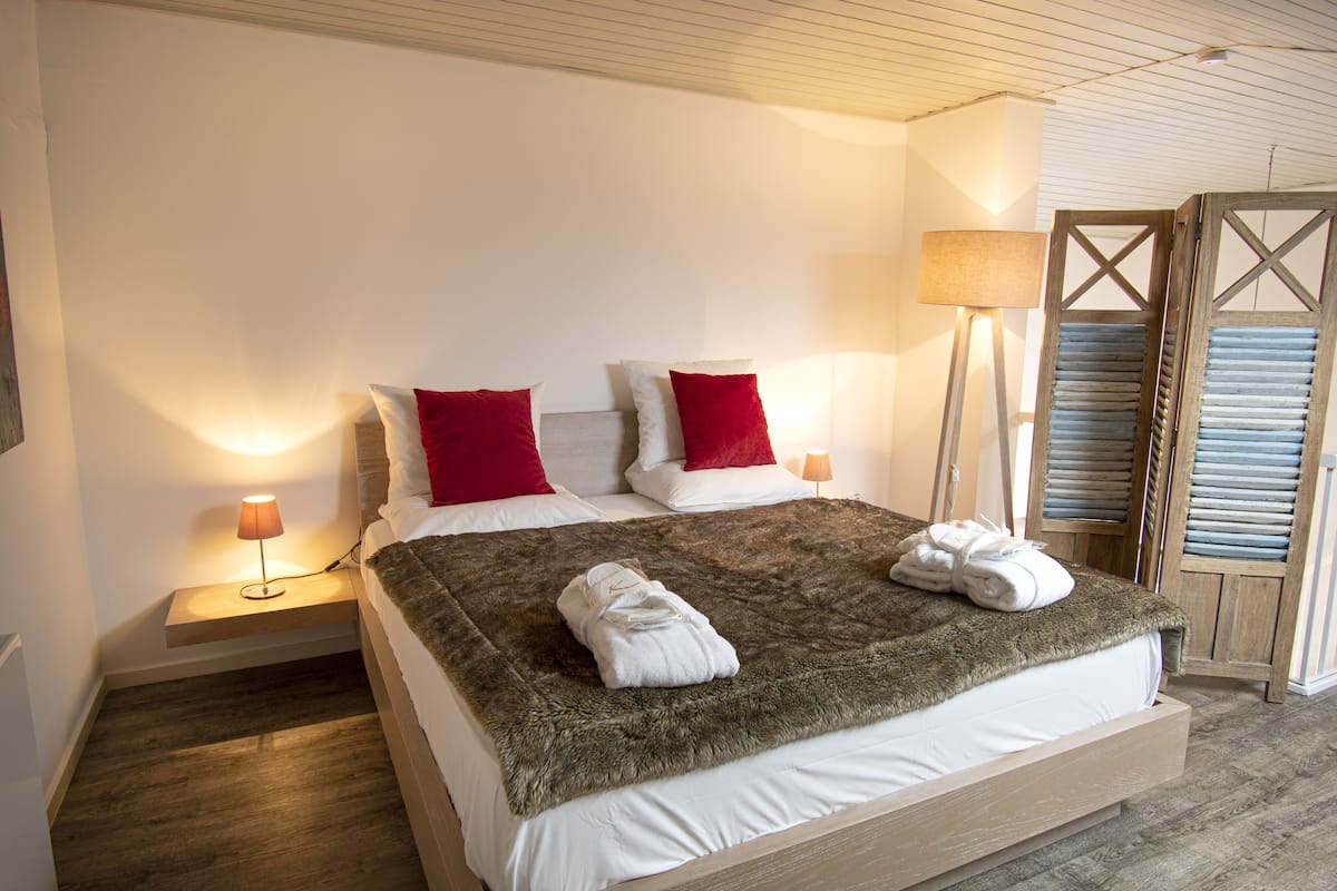 Cama bem arrumada com um cobertor fofo, duas almofadas vermelhas e um par de toalhas sobre a cama. Há uma mesinha de cabeceira com uma luminária e uma prateleira fixada na parede.