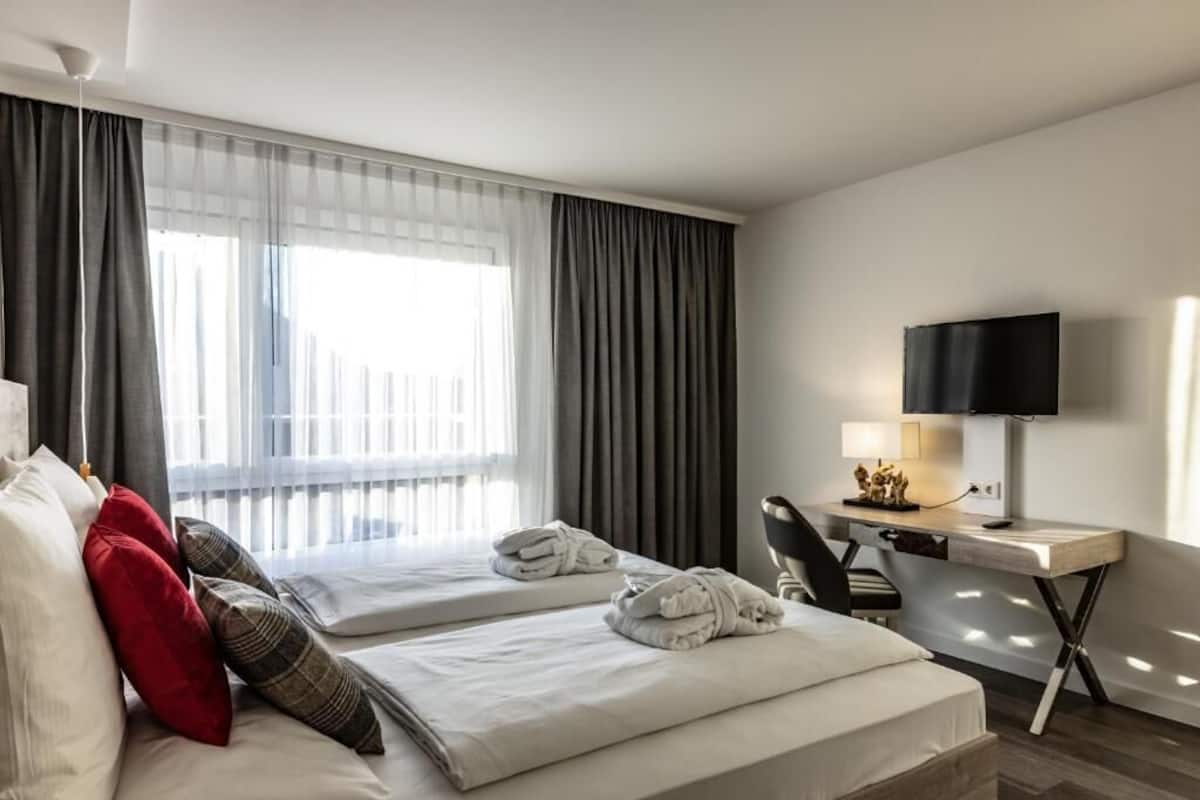 Quarto de hotel com duas camas, uma mesa, uma televisão e uma janela com cortinas.