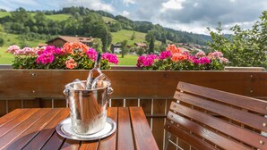 A wooden table with a metal pitcher and a glass of water, set against a backdrop of colorful flowers and a scenic view of green hills and houses. - Karma Bavaria (Schliersee)