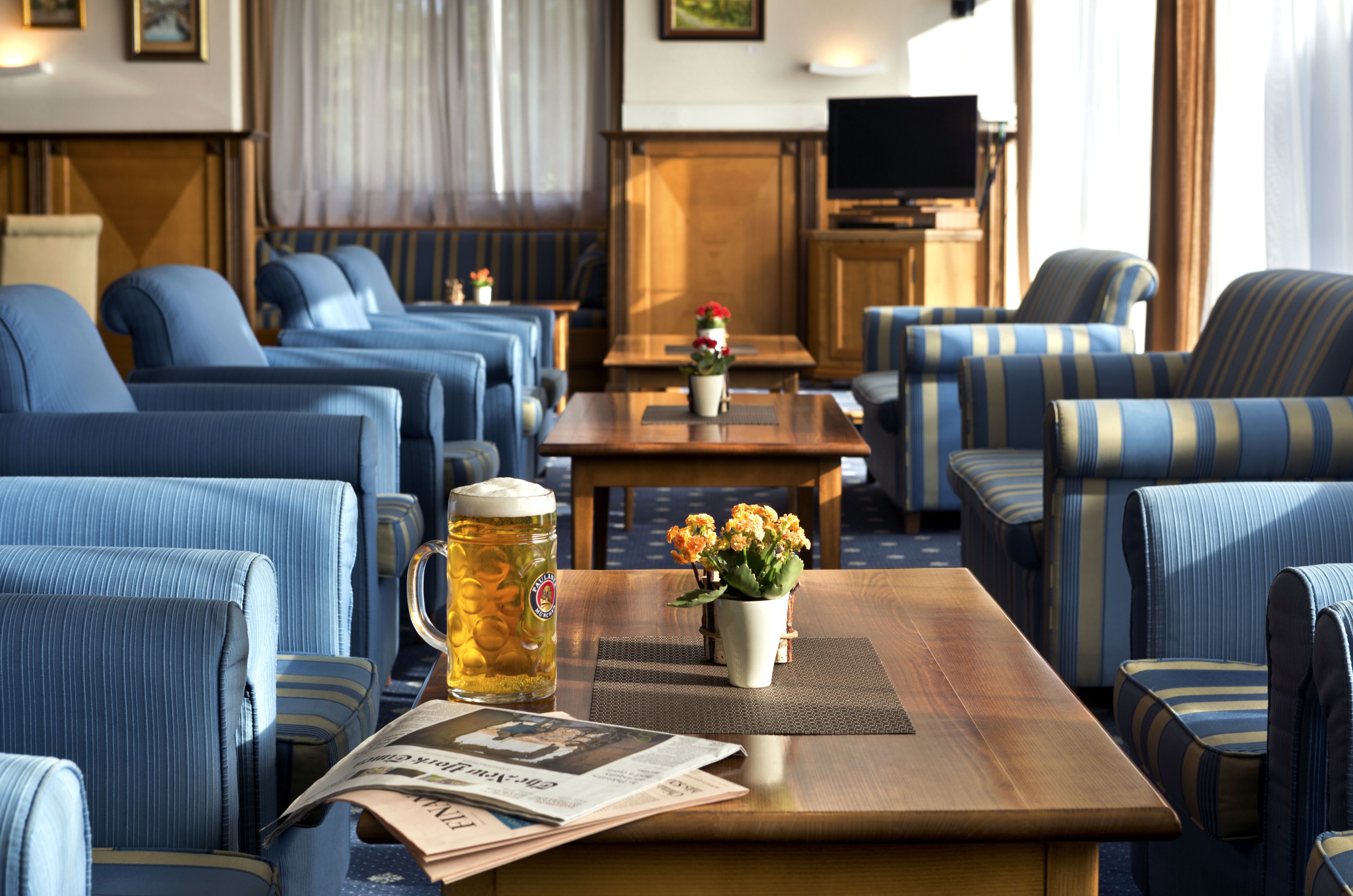 Un vestíbulo de hotel con sofás azul rayados, una mesa de centro de madera con un periódico, un vaso de cerveza y un jarrón con flores.