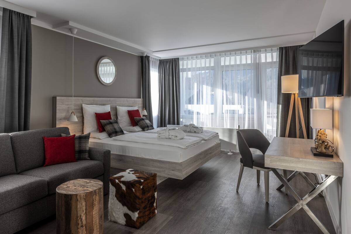 Quarto de hotel moderno com cama, sofá, escrivaninha e espelho redondo.
