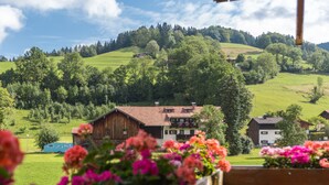 A rural landscape with a wooden house, a blue trailer, and a white building in the background. - Karma Bavaria (Schliersee)
