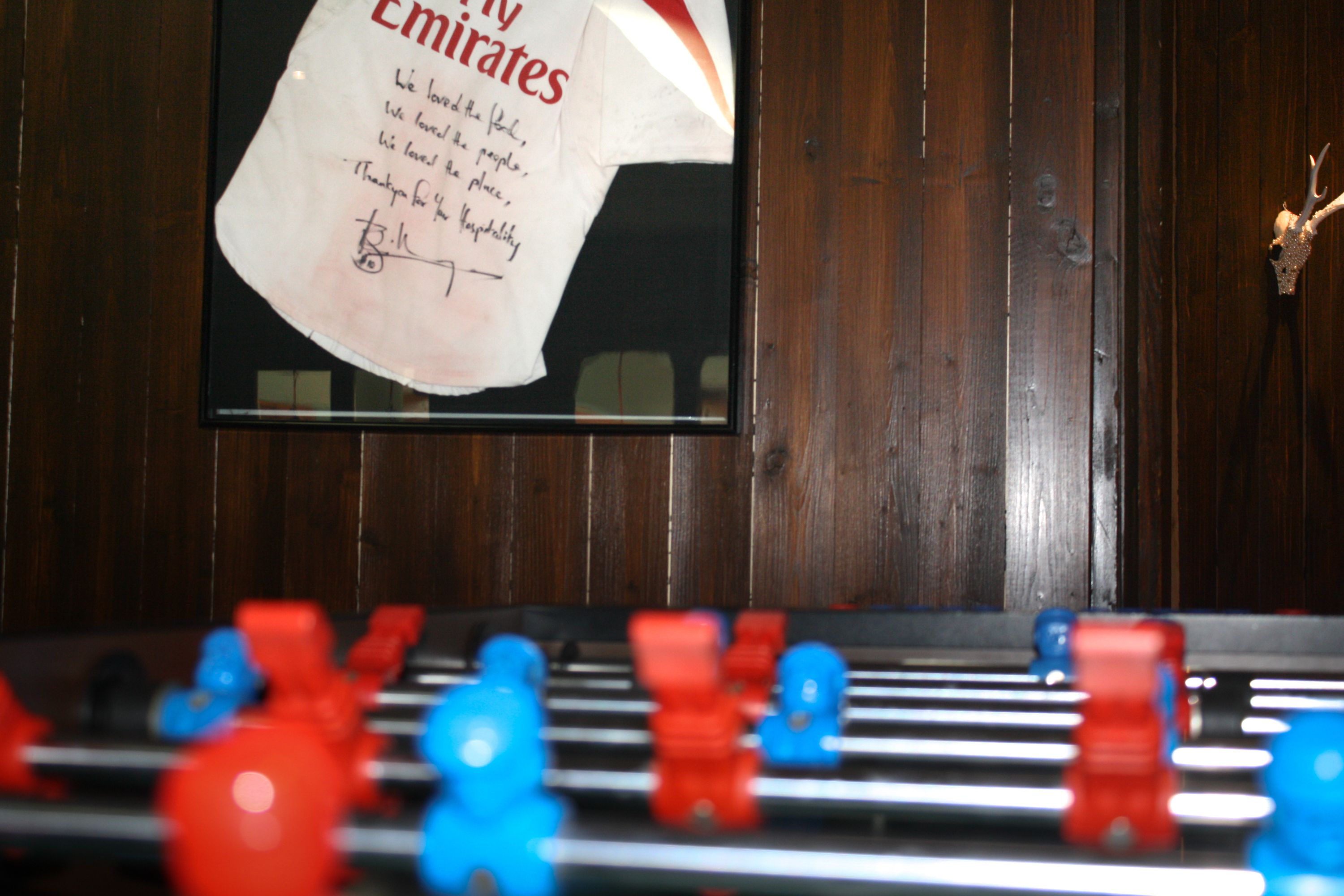 Une table de baby-foot avec des figurines rouges et bleues, un panneau encadré avec du texte, et un lambris en bois en arrière-plan.