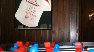 Une table de baby-foot avec des figurines rouges et bleues, un panneau encadré avec du texte, et un lambris en bois en arrière-plan.