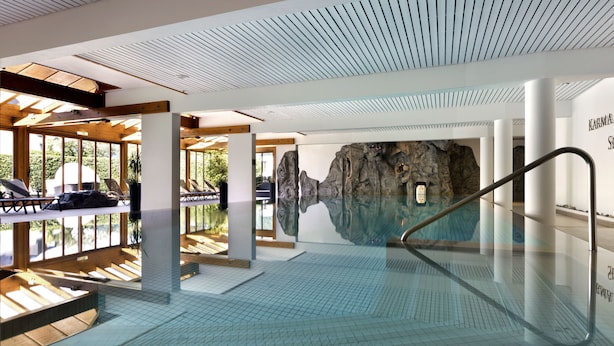 A modern indoor swimming pool with a glass wall, wooden beams, and a view of a rocky landscape. - Karma Bavaria (Schliersee)