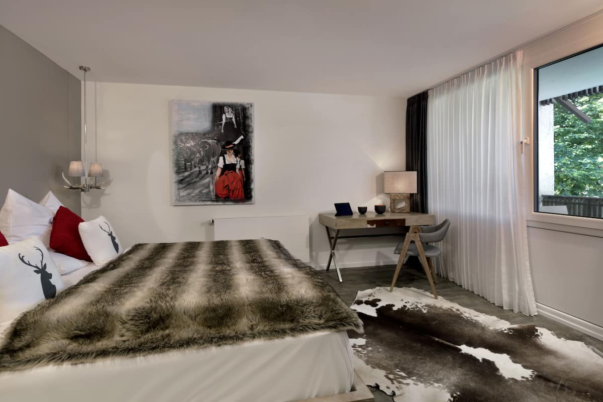 Quarto de hotel com cama, escrivaninha, cadeira, luminária, pintura e uma grande janela com cortinas.