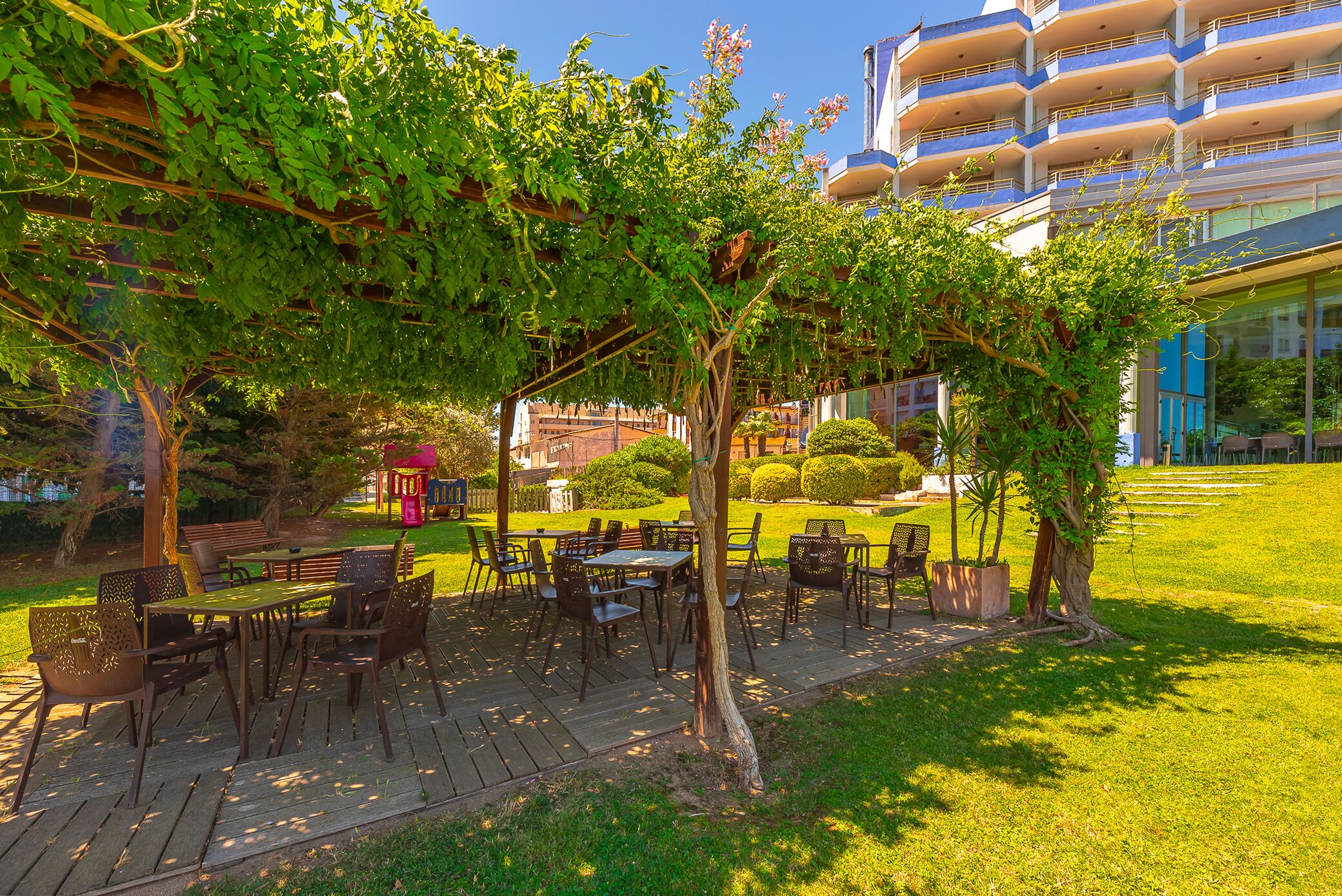 An outdoor patio with tables and chairs, surrounded by greenery and a building in the background.