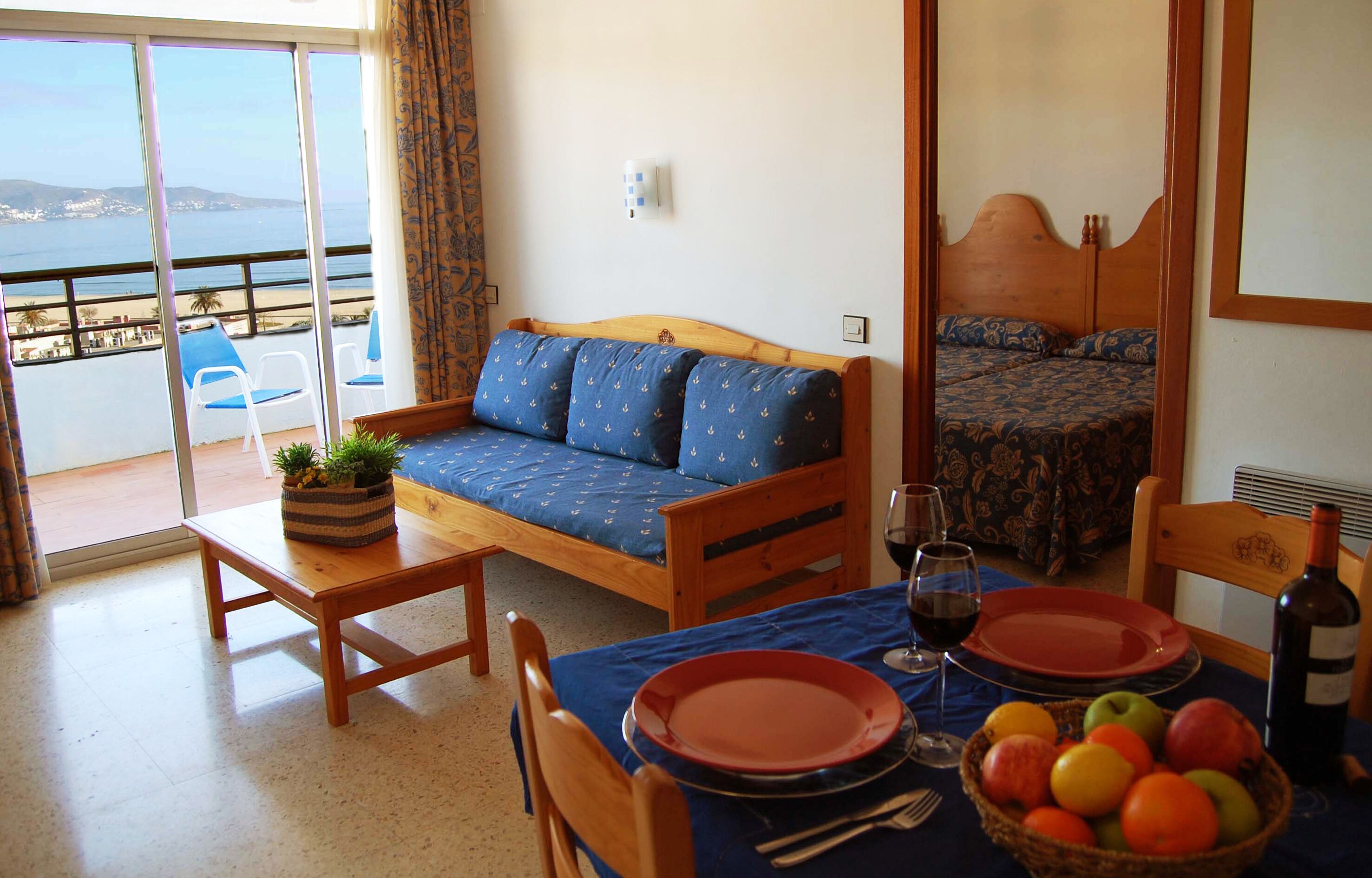 Una habitación con balcón, sofá, cama, mesa de comedor servida con platos y una botella de vino, y una cesta de frutas.