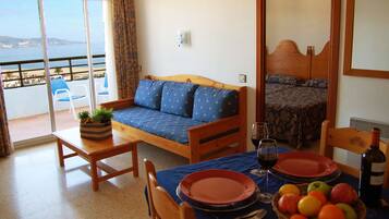 Una habitación con balcón, sofá, cama, mesa de comedor servida con platos y una botella de vino, y una cesta de frutas.