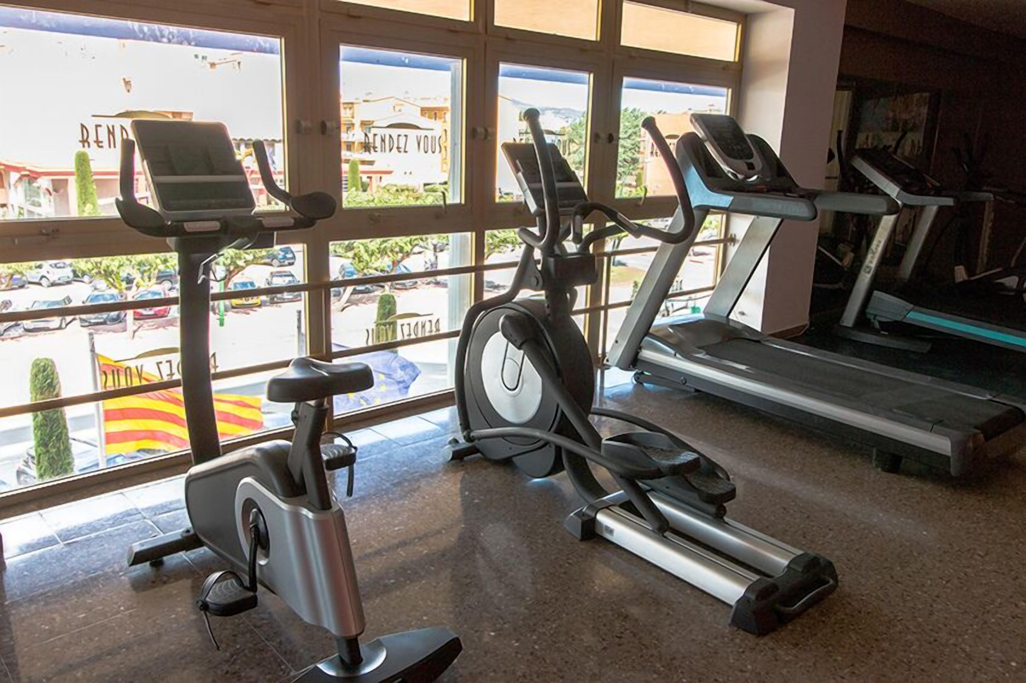 Une salle de sport équipée de vélos d’appartement, de tapis de course et d’une fenêtre donnant sur des voitures et des immeubles en contrebas.