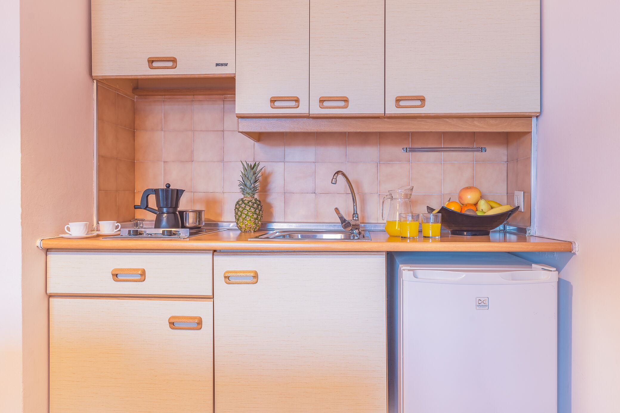 Une cuisine compacte avec des meubles en bois, un évier, une machine à café, un panier de fruits et un petit réfrigérateur.