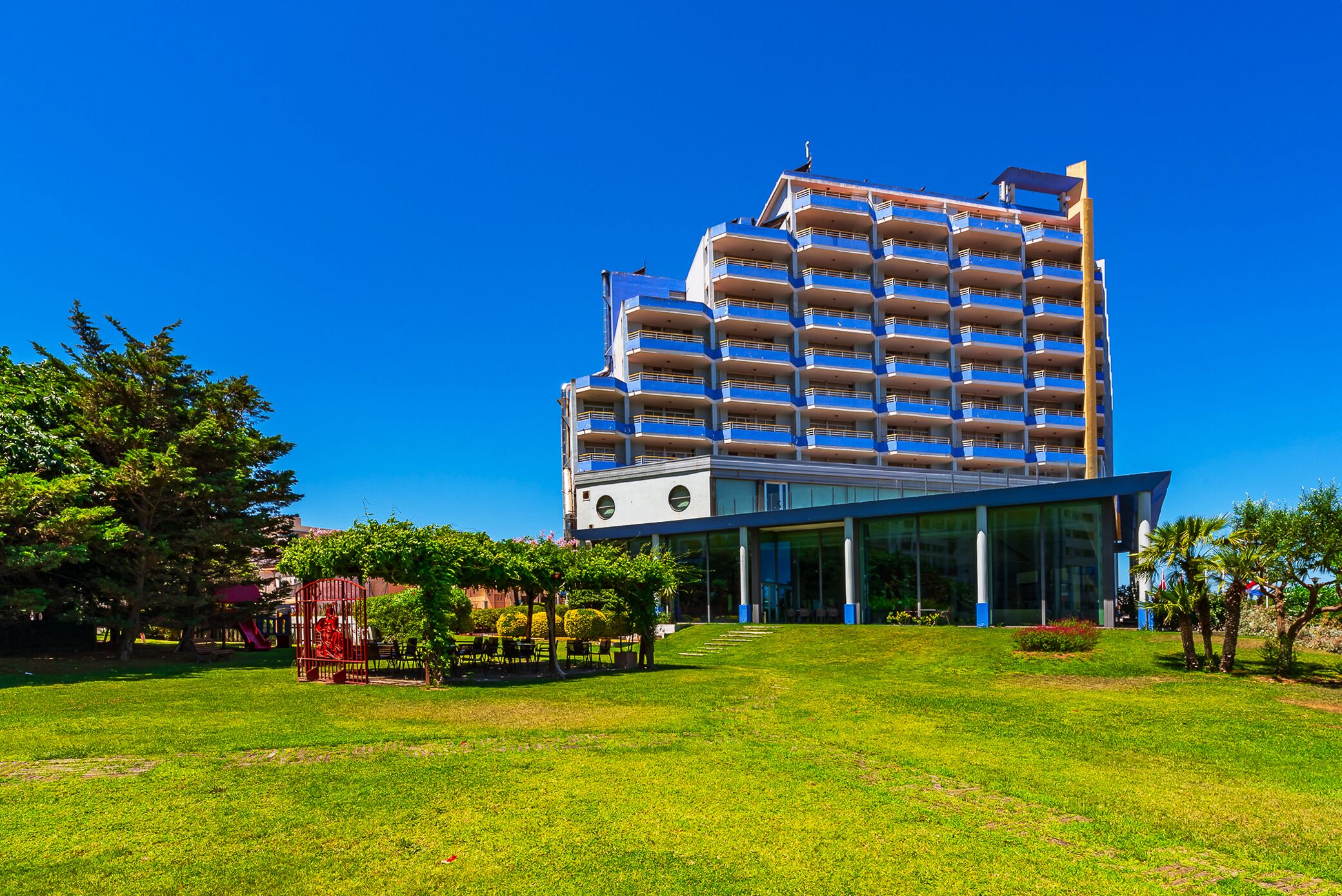 A modern hotel with a glass facade, multiple balconies, and a well-maintained lawn.