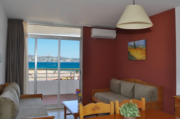 A room with a sofa, a dining table, and a view of the beach and sea through a large window. - Xon's Platja ApartHotel (Castello d'Empuries)