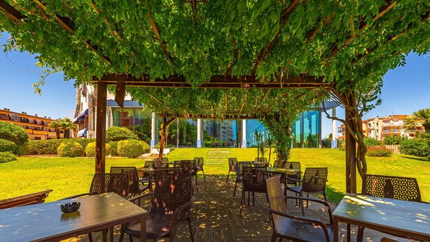 Un espace extérieur aménagé en terrasse, avec des tables et des chaises sous une pergola, offrant une vue sur une pelouse bien entretenue et des bâtiments en arrière-plan.