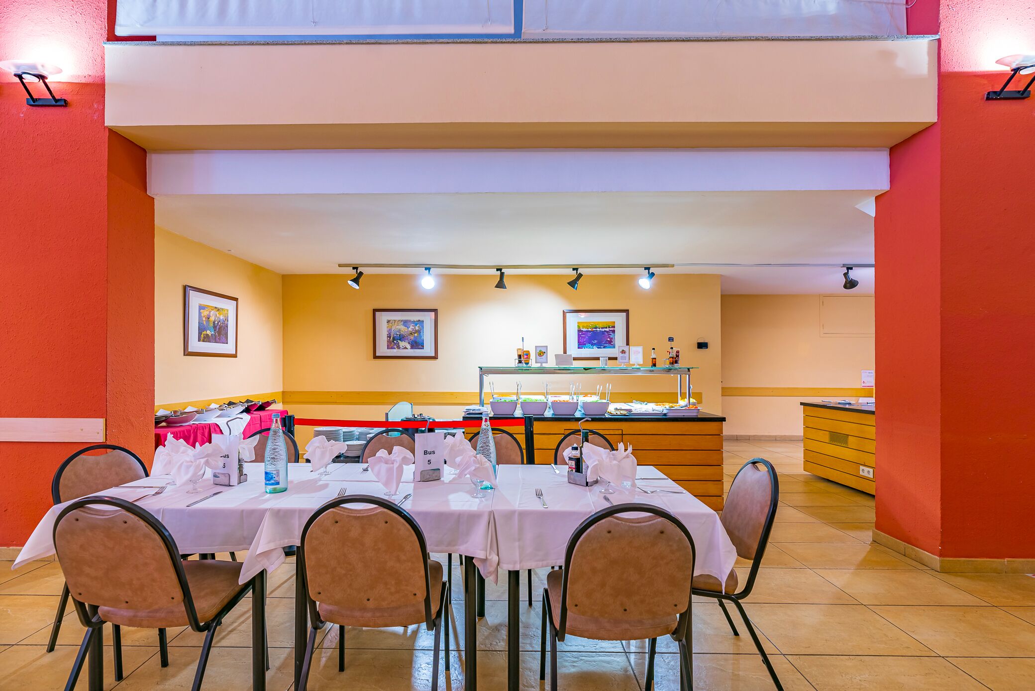 Un comedor con mesas dispuestas para el servicio, un mostrador de buffet y una cocina al fondo.