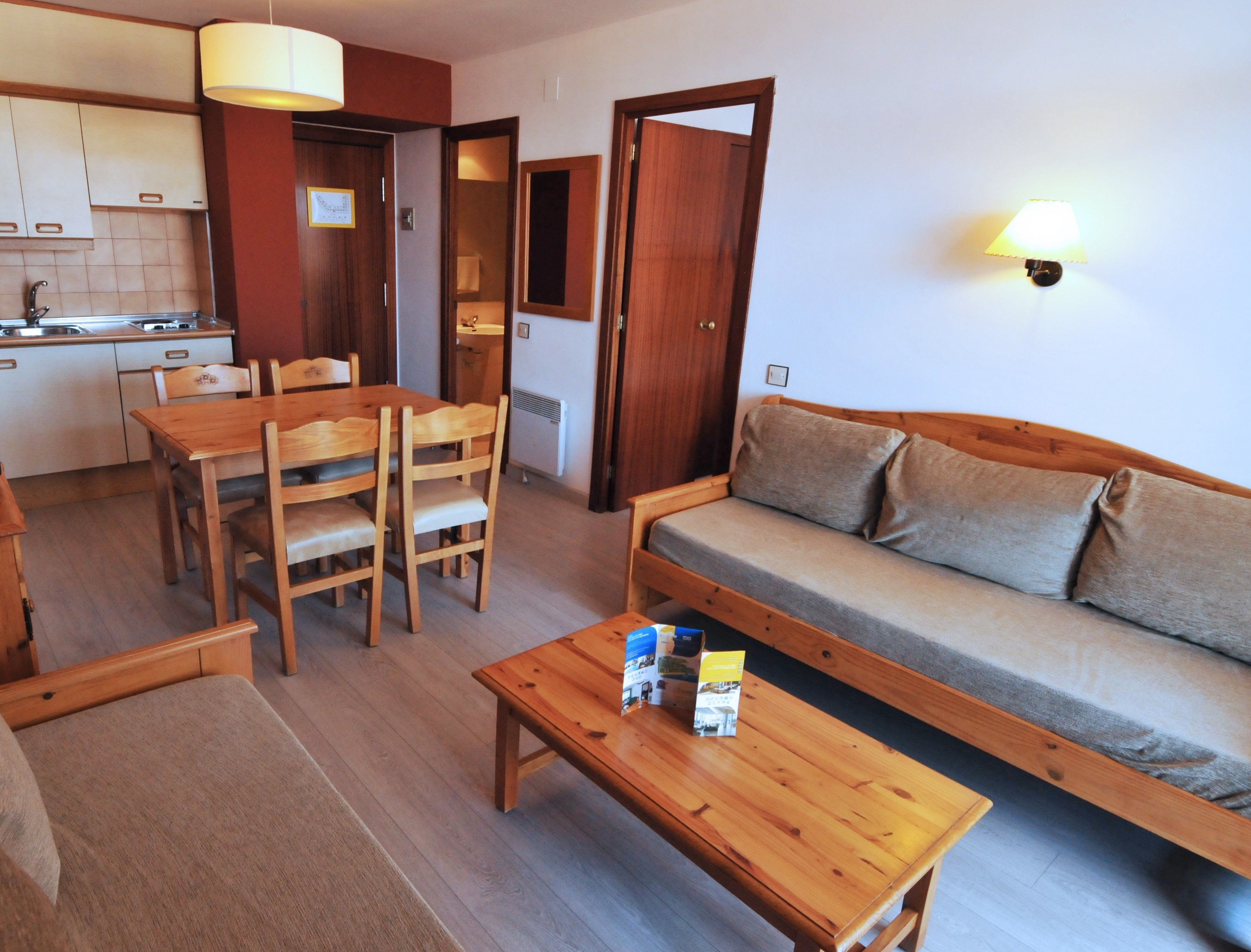 A compact living space with a wooden dining table, chairs, a sofa, and a small coffee table. A kitchen area with cabinets and a sink is visible in the background.