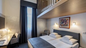 A hotel room with a bed, desk, chair, artwork, and a view of the city.