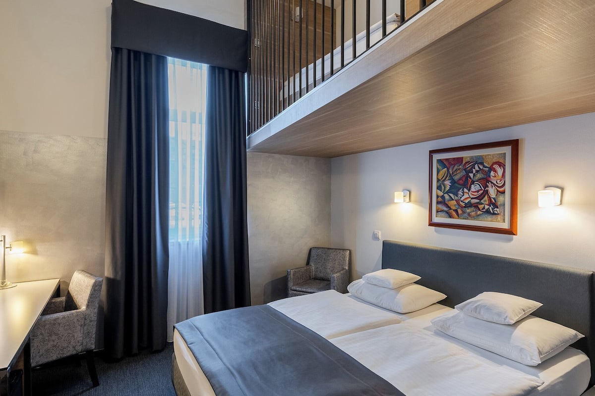 A hotel room with a bed, desk, chair, artwork, and a view of the city.