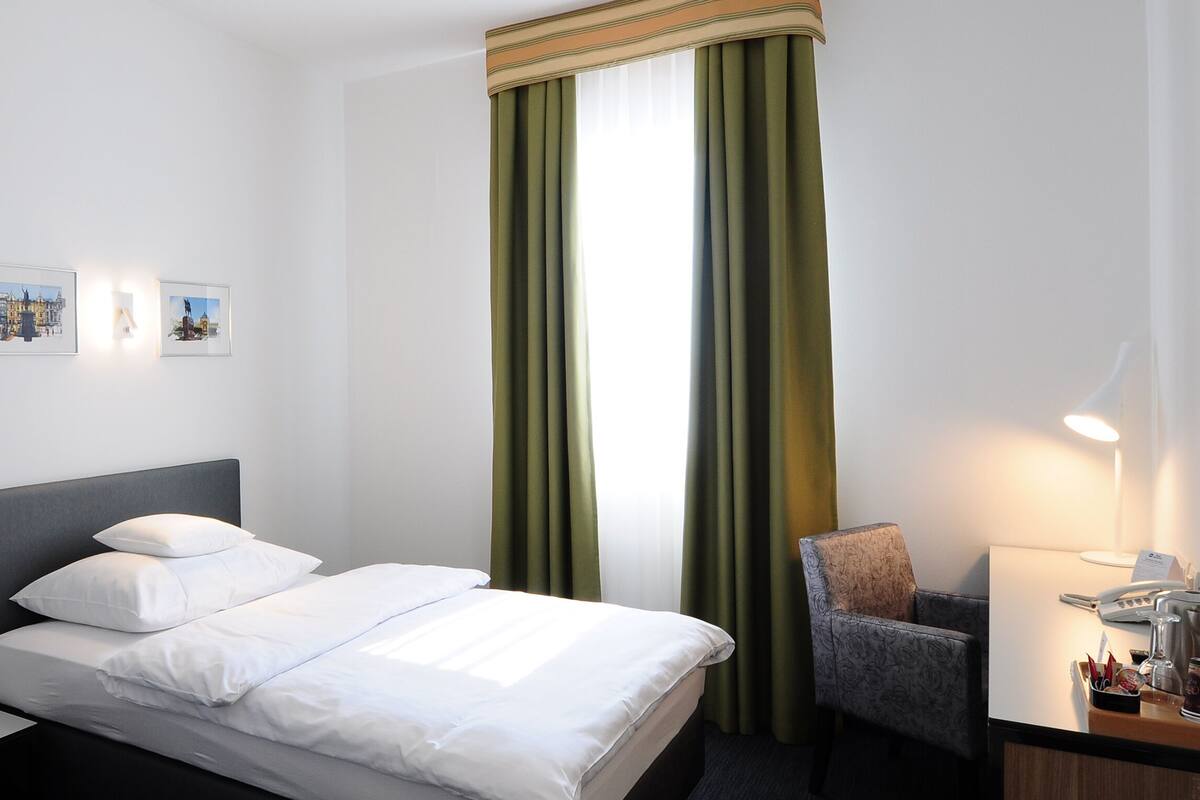 A hotel room with a bed, a desk with a telephone and coffee maker, a chair, and two framed pictures on the wall.