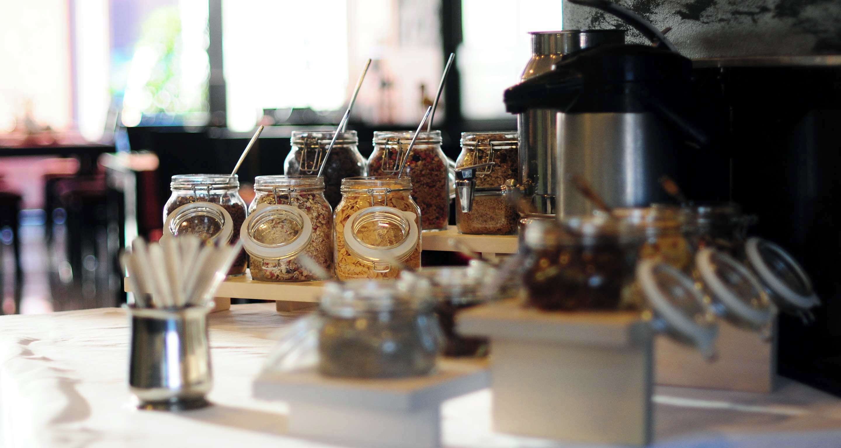 Esposizione di diversi alimenti in vasetti con cucchiaini, una macchina del caffè e un piccolo contenitore metallico con utensili.
