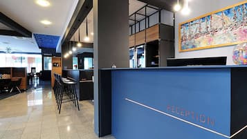 A modern reception area with a blue reception desk labeled "RECEPTION," a bar area, and a terrace visible in the background.