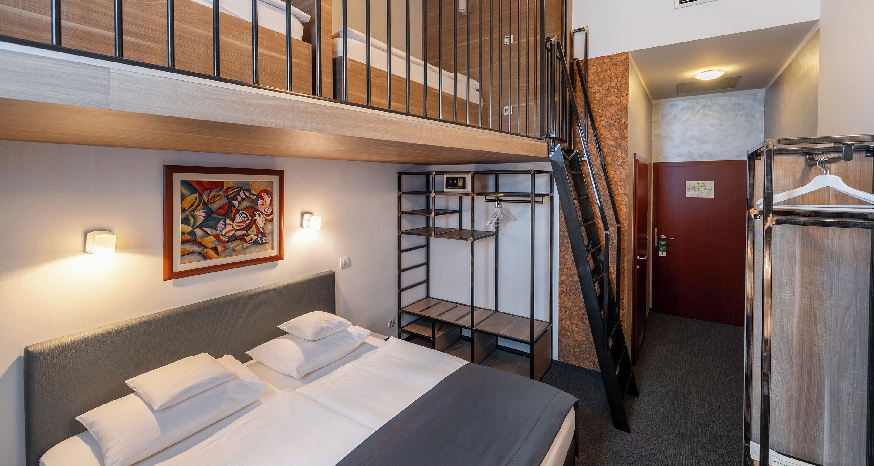 Quarto de hotel com beliche, um quadro, um guarda-roupa e uma escada.
