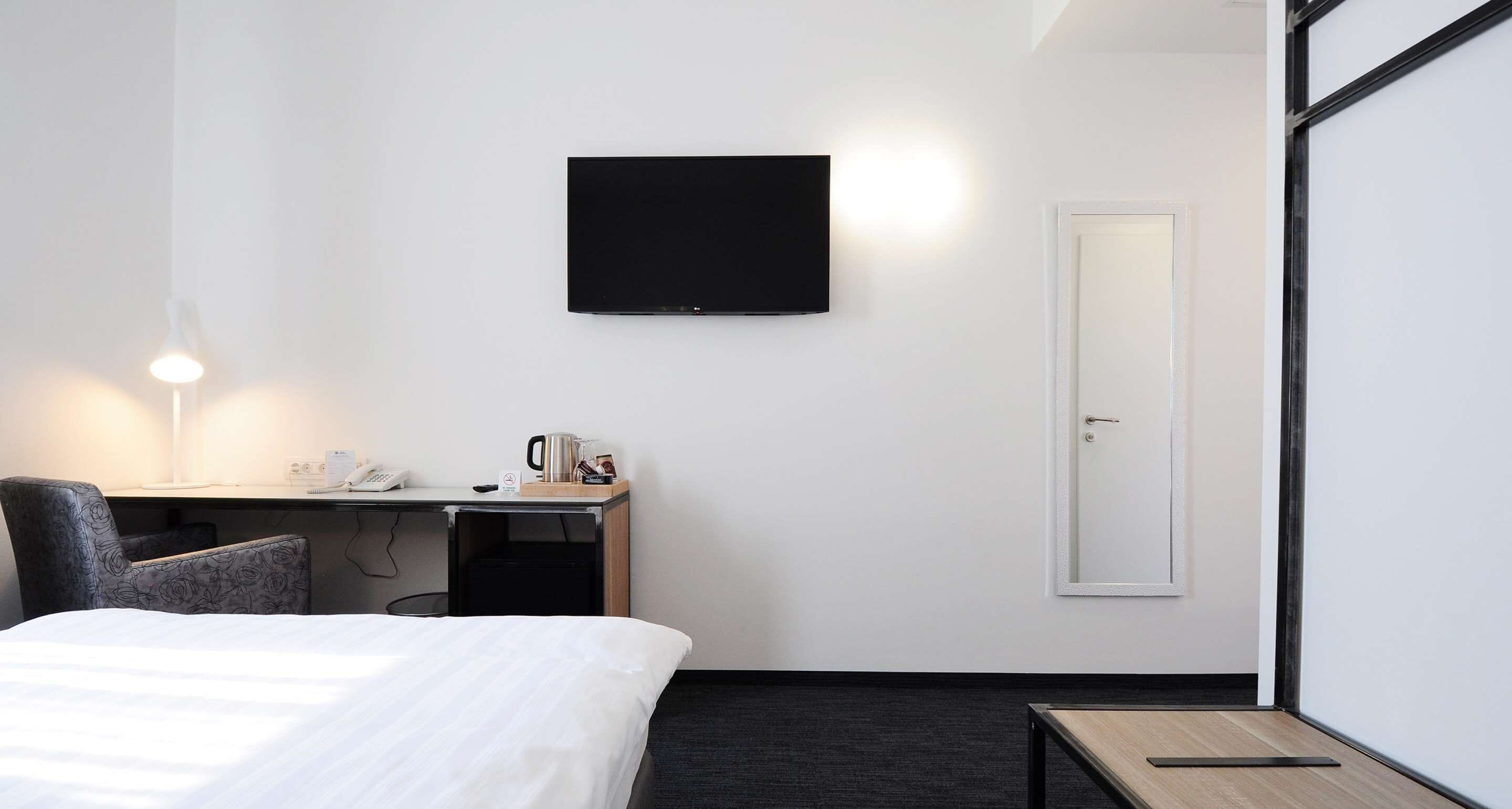 A hotel room with a bed, a desk with a chair, a flat-screen TV, a door, and a small table.