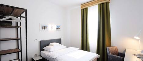 Habitación de hotel con cama, una silla, escritorio, estante y ventana con cortinas.