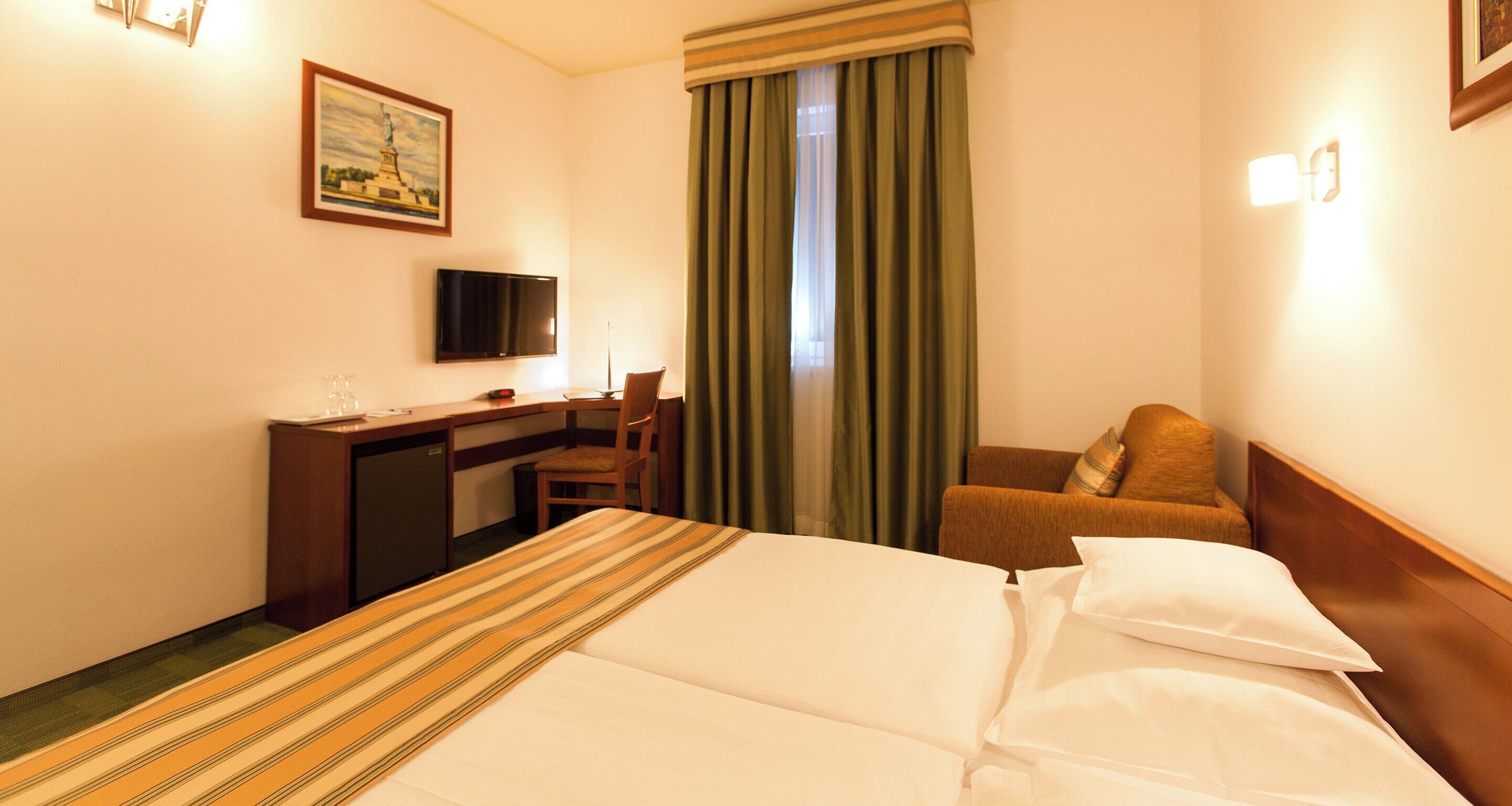 Habitación de hotel con cama, escritorio, silla, televisor y ventana con cortinas.