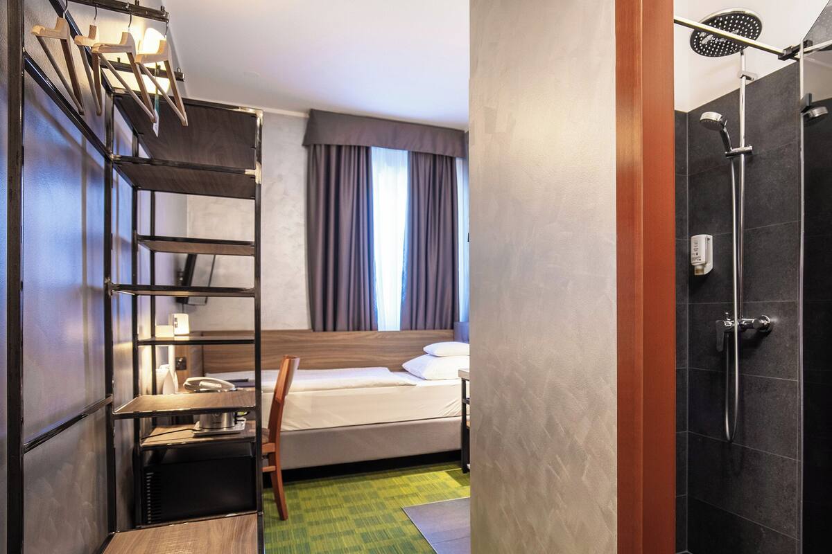 A hotel room with a bed, a desk, a chair, a shower, and a towel rack.