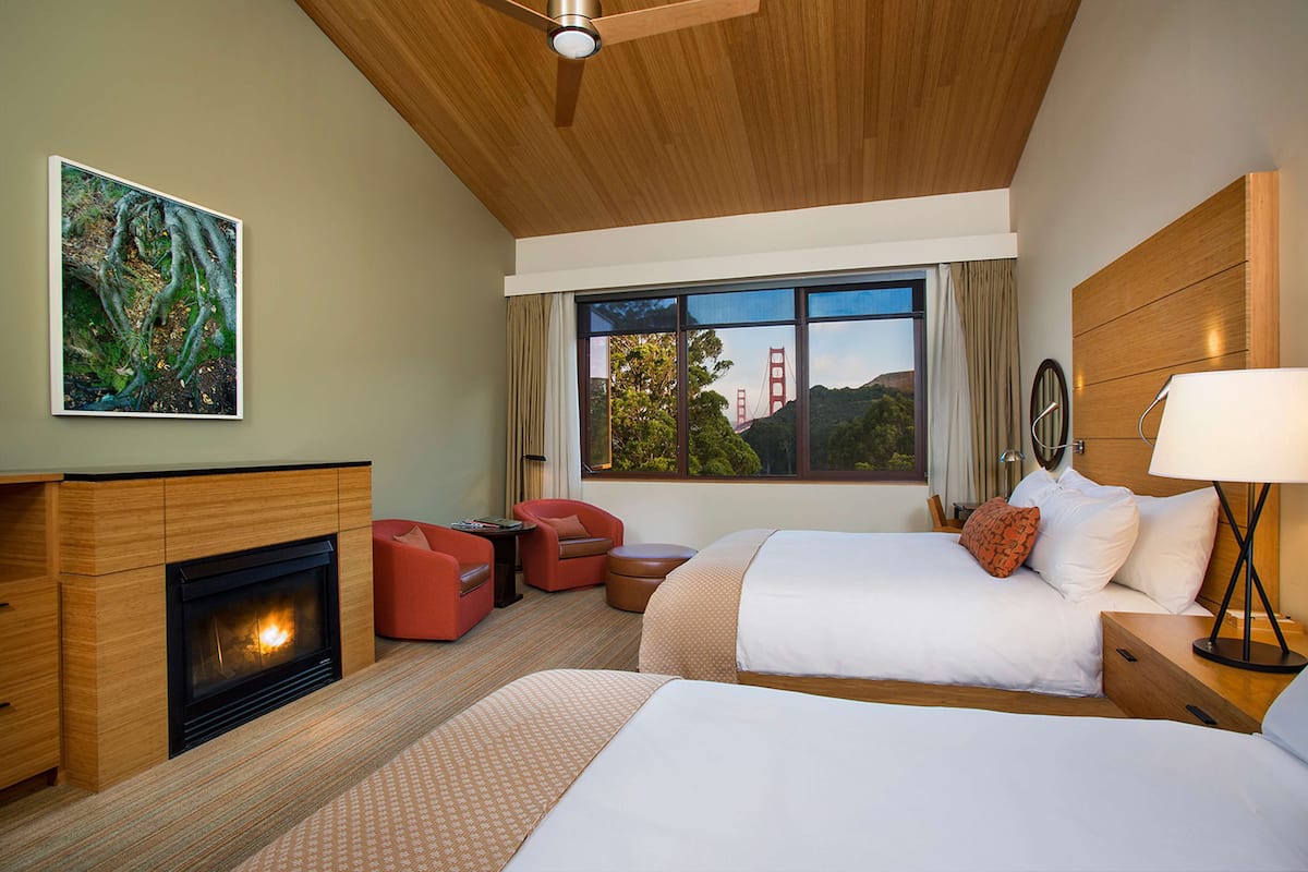 A hotel room with a fireplace, two beds, a red armchair, a wooden headboard, and a large window with a view of the Golden Gate Bridge.