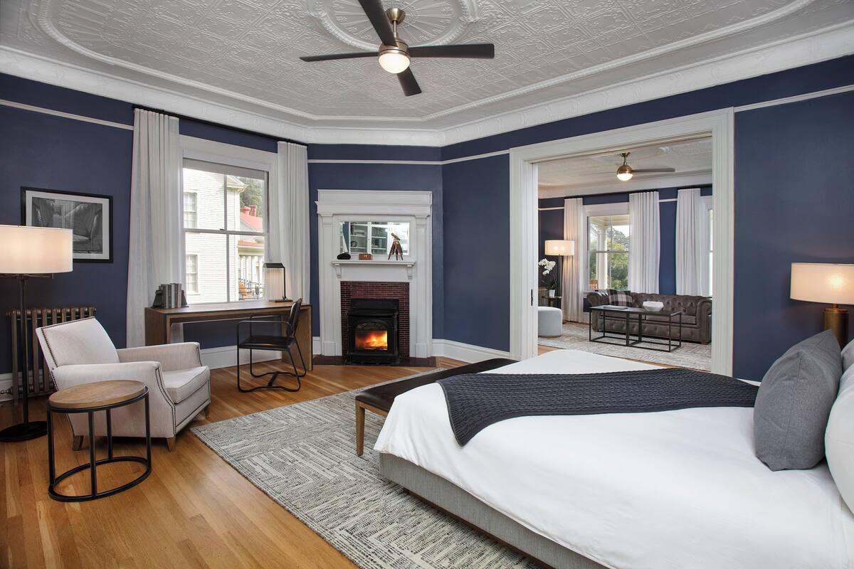 A bedroom with a large bed, a desk, a chair, a fireplace, and a view of the outside.