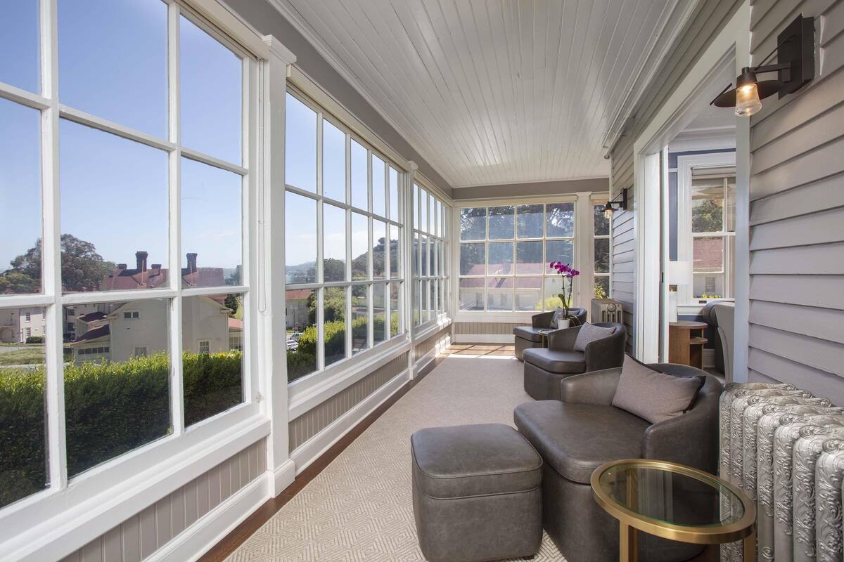 A sunroom with large windows, a gray sofa, a glass table, and a view of the neighborhood.