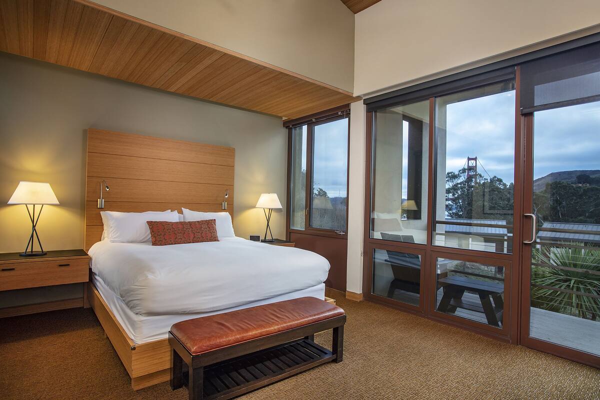 A modern hotel room with a large bed, two bedside lamps, a wooden headboard, and a balcony with a view of a bridge.